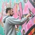 A young guy in a gray hoodie paints graffiti in pink and green c