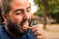 Young guy with long beard eats ice cream with a plastic spoon in the park