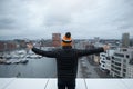 A young guy look at city from the roof of building with his arms raised. Black jacket and orange cap. Belgium, Antwerp.