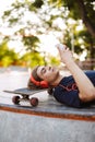 Young guy in orange headphones lying on skateboard dreamily using cellphone while spending time at skatepark Royalty Free Stock Photo