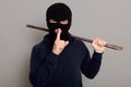 Young guy robber man dressed in a black hoodie with masked face, holds iron crowbar in his hands, shows silence gesture, keeping