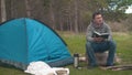 A young guy sits on a large log near a blue tent and Temos in the forest and reads a book.