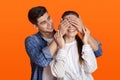 Young guy stands behind lady and closes eyes of smiling woman isolated on orange background