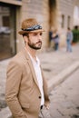 A young guy with a straw hat, a white unbuttoned shirt and a sand jacket, a small beard walks around the city of
