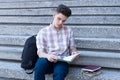 Young guy student sitting on the stairs of a school, college or university and reading a precis. Student preparing for exam