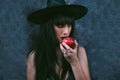 Young Halloween witch woman portrait eating a red apple. Beauty Angry Vampire Witch lady with black mouth in the darkness, wearing