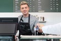 Young handsom male barista wearing apron, getting paid by credit card payment from customer while standing behind coffee shop