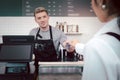 Young handsom male barista wearing apron, getting paid by credit card payment from customer while standing behind coffee shop