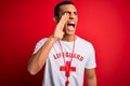 Young handsome african american lifeguard man wearing t-shirt with red cross and whistle shouting and screaming loud to side with Royalty Free Stock Photo