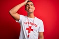 Young handsome african american lifeguard man wearing t-shirt with red cross and whistle smiling confident touching hair with hand