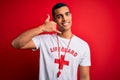 Young handsome african american lifeguard man wearing t-shirt with red cross and whistle smiling doing phone gesture with hand and