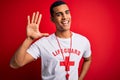 Young handsome african american lifeguard man wearing t-shirt with red cross and whistle Waiving saying hello happy and smiling,