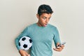 Young handsome african american man holding football ball looking at smartphone afraid and shocked with surprise and amazed Royalty Free Stock Photo