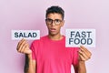 Young handsome african american man holding salad and fast food message paper in shock face, looking skeptical and sarcastic, Royalty Free Stock Photo