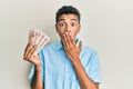 Young handsome african american man holding 10 united kingdom pounds banknotes covering mouth with hand, shocked and afraid for