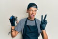 Young handsome african american man tattoo artist wearing professional uniform and gloves holding tattooer machine smiling with Royalty Free Stock Photo