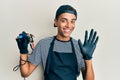 Young handsome african american man tattoo artist wearing professional uniform and gloves holding tattooer machine waiving saying Royalty Free Stock Photo