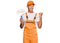 Young handsome african american man wearing cap and painter clothes holding painting roll pointing thumb up to the side smiling Royalty Free Stock Photo