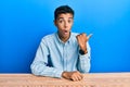 Young handsome african american man wearing casual clothes sitting on the table surprised pointing with hand finger to the side, Royalty Free Stock Photo