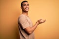 Young handsome african american man wearing casual t-shirt standing over yellow background pointing aside with hands open palms Royalty Free Stock Photo