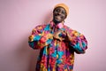 Young handsome african american man wearing colorful coat and cap over pink background smiling in love doing heart symbol shape Royalty Free Stock Photo
