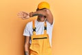 Young handsome african american man wearing handyman uniform over yellow background covering eyes with arm, looking serious and
