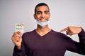 Young handsome african american man wearing medical mask holding virus alert reminder with surprise face pointing finger to Royalty Free Stock Photo