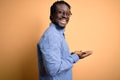 Young handsome african american man wearing shirt and glasses over yellow background pointing aside with hands open palms showing Royalty Free Stock Photo