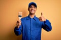 Young handsome african american painter man wearing uniform using painting brush happy with big smile doing ok sign, thumb up with