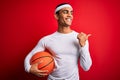 Young handsome african american sportsman holding basketball ball over red background pointing and showing with thumb up to the Royalty Free Stock Photo