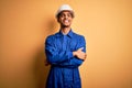 Young handsome african american worker man wearing blue uniform and security helmet happy face smiling with crossed arms looking Royalty Free Stock Photo