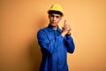 Young handsome african american worker man wearing blue uniform and security helmet Holding symbolic gun with hand gesture,