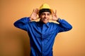 Young handsome african american worker man wearing blue uniform and security helmet Trying to hear both hands on ear gesture, Royalty Free Stock Photo