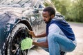 Young handsome African guy holding green sponge, washing car wheel with foam. Cleaning of modern rims of luxury blue Royalty Free Stock Photo