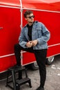 Young handsome american hipster man in stylish black pants in a t-shirt in a blue trendy denim jacket posing near a red vintage Royalty Free Stock Photo