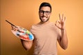 Young handsome artist man with beard painting using bursh and palette with colors doing ok sign with fingers, excellent symbol