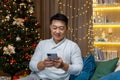 Young handsome Asian man sitting at home near the Christmas tree. Holding a mobile phone, typing Royalty Free Stock Photo