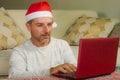 Young handsome and attractive happy man in Santa hat at home online shopping on internet with laptop computer using credit card bu Royalty Free Stock Photo