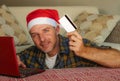 Young handsome and attractive happy man in Santa hat at home online shopping on internet with laptop computer using credit card bu Royalty Free Stock Photo