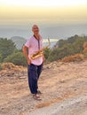 Young handsome and bald male saxophonist stands against the sunset with a golden saxophone musical instrument and smiles. In a Royalty Free Stock Photo