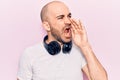 Young handsome bald man listening to music using headphones shouting and screaming loud to side with hand on mouth Royalty Free Stock Photo