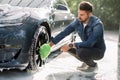 Young handsome bearded guy holding green sponge, washing car wheel with foam. Cleaning of modern rims of luxury blue Royalty Free Stock Photo