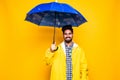 Young handsome bearded indian man in yellow raincoat with blue umbrella cover from rain isolated over orange background Royalty Free Stock Photo