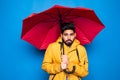 Young handsome bearded indian man in yellow raincoat with red umbrella cover from rain get cold isolated over blue background Royalty Free Stock Photo