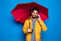 Young handsome bearded indian man in yellow raincoat with red umbrella cover from rain talking in the phone isolated over blue Royalty Free Stock Photo