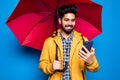 Young handsome bearded indian man in yellow raincoat with red umbrella cover from rain use phone isolated over blue background Royalty Free Stock Photo