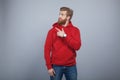 Young handsome bearded man wearing red sweatshirt standing on the grey background and pointing his finger on the left