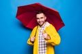 Young handsome bearded man in yellow raincoat with red umbrella pointed on you isolated over blue background Royalty Free Stock Photo