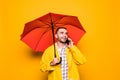 Young handsome bearded man in yellow raincoat with red umbrella talking by mobile phone isolated over orange background Royalty Free Stock Photo