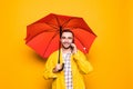 Young handsome bearded man in yellow raincoat with red umbrella talking by mobile phone isolated over orange background Royalty Free Stock Photo